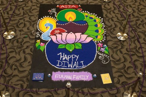 An artistic representation of a peacock, candle and lotus made of colored sand. The art says "Happy Diwali!"